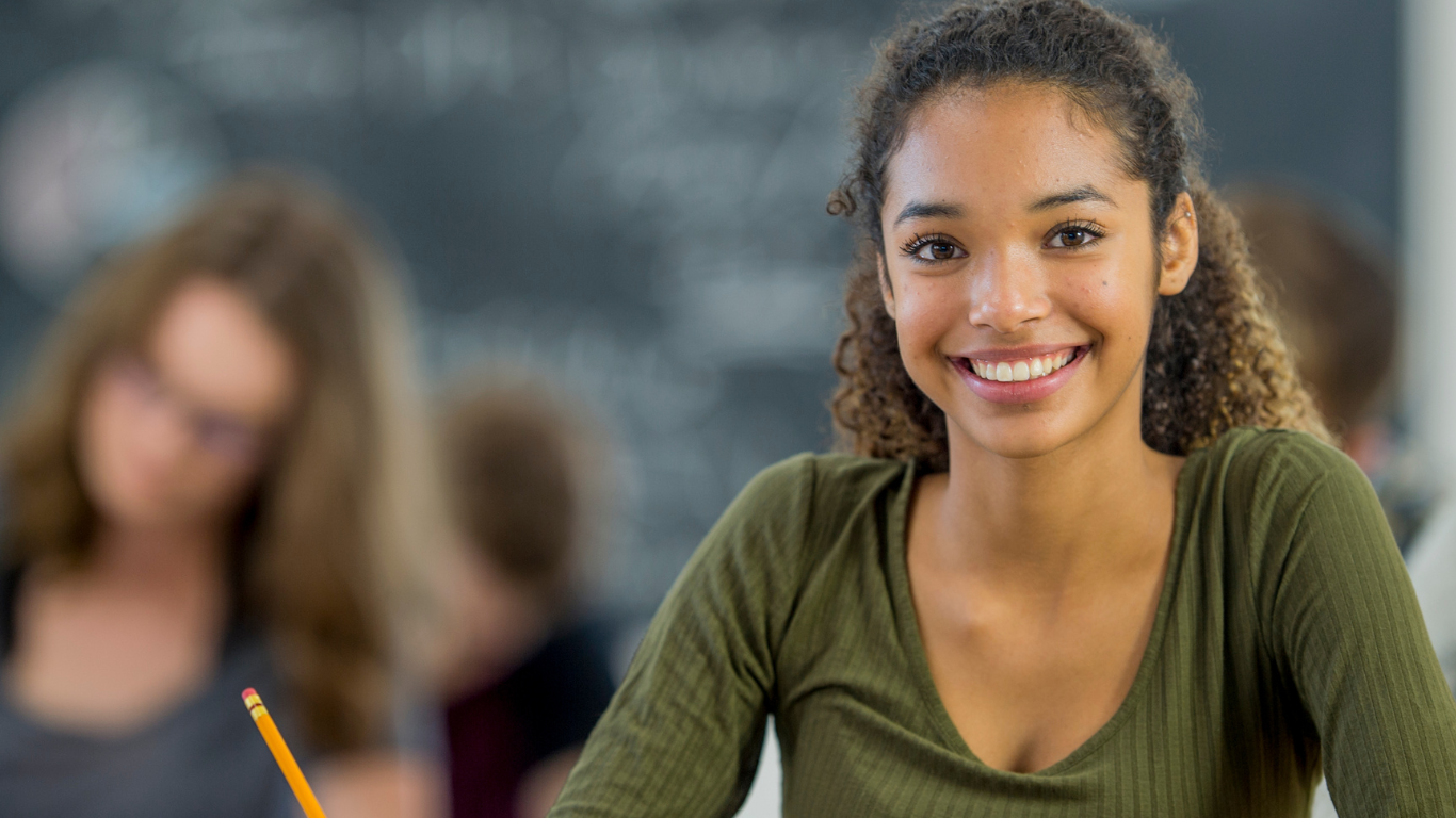 A smiling person sitting at a desk in front of a blackboard